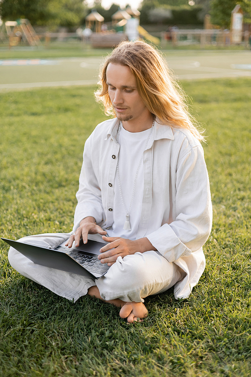 young long haired man in white cotton clothes sitting in easy pose and using laptop on green lawn