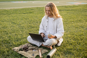 young man in white clothes holding puer tea and using laptop while sitting near thermos and ceramic teapot on green lawn