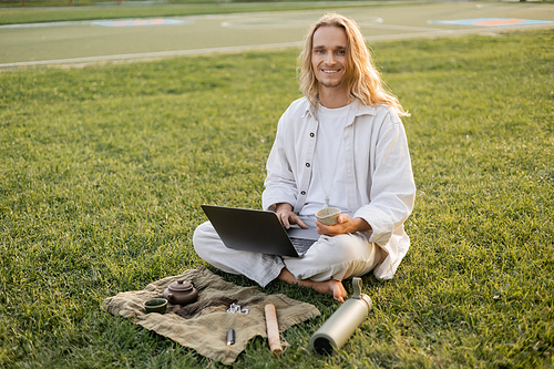 happy long haired man sitting with laptop and clay cup near thermos and linen rug with teapot on grassy lawn