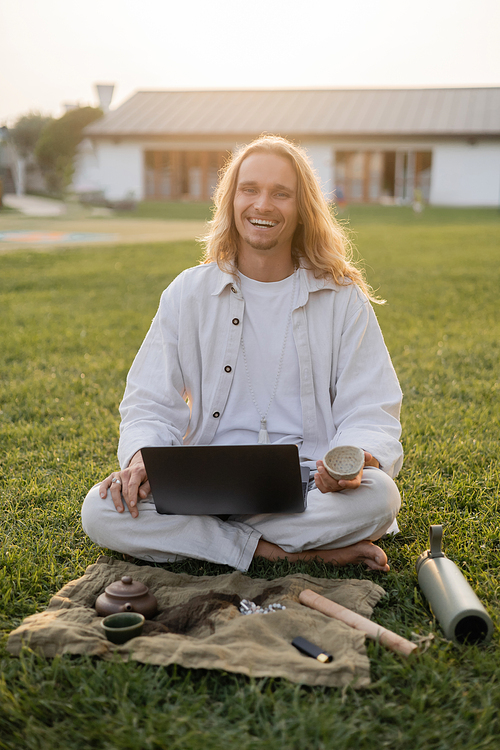 carefree man smiling at camera while sitting on green grass with laptop and clay cup near thermos and linen rug with teapot