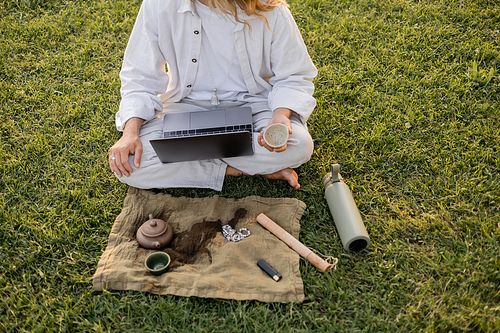 partial view of yoga man sitting on green lawn with laptop and puer tea near thermos and linen rug with clay teapot and mala beads
