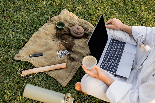 partial view of man with laptop holding clay cup near teapot and mala beads on linen rug on green lawn