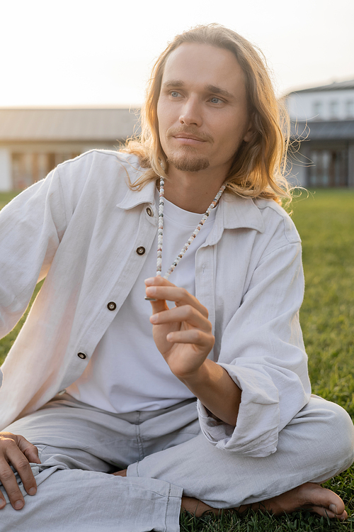 long haired man in linen clothes touching mala beads and looking away during meditation outdoors