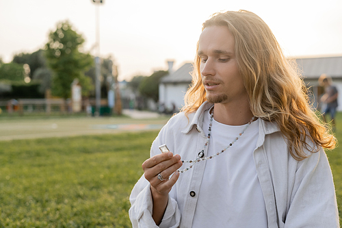 thoughtful yoga man in white linen shirt looking at crystal on mala beads while standing in countryside outdoors