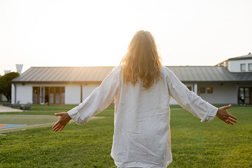 back view of long haired man in white linen shirt meditating with outstretched hands near cottage outdoors