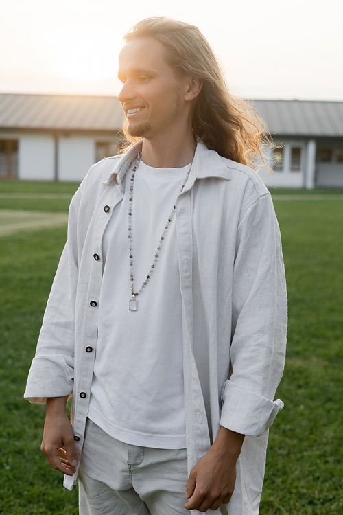 young long haired yoga man in linen shirt and beads smiling while standing in countryside outdoors