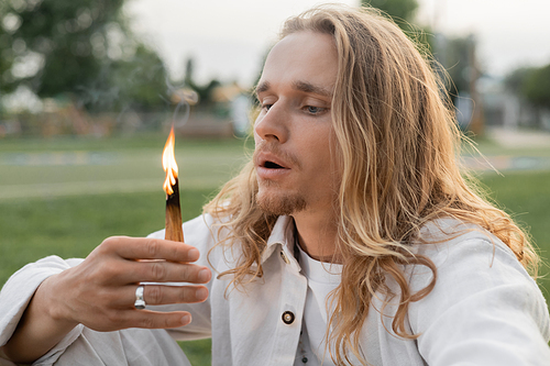 long haired man in white clothes blowing at burning palo santo stick