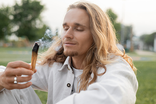 pleased man with closed eyes holding smoldering palo santo stick during spiritual ritual outdoors