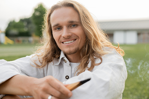 carefree long haired man holding fragrant palo santo stick and looking at camera outdoors