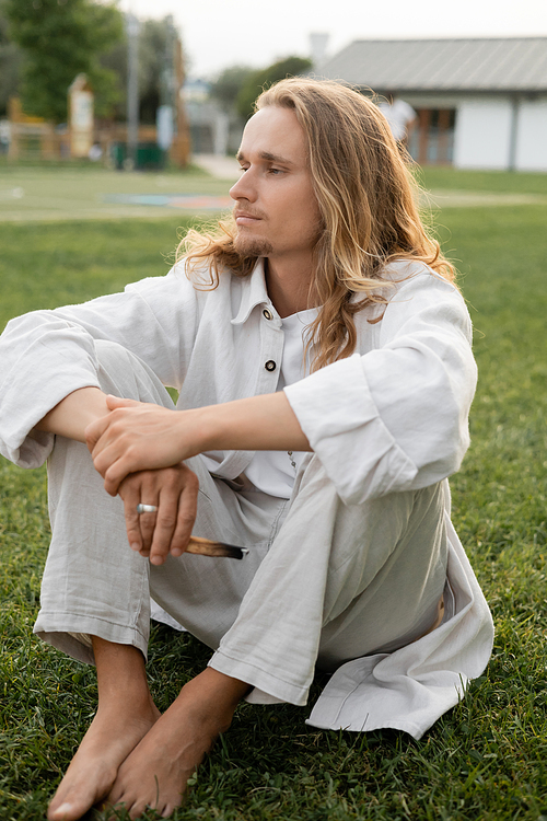 calm barefoot man in white linen clothes looking away while sitting with aromatic palo santo stick outdoors