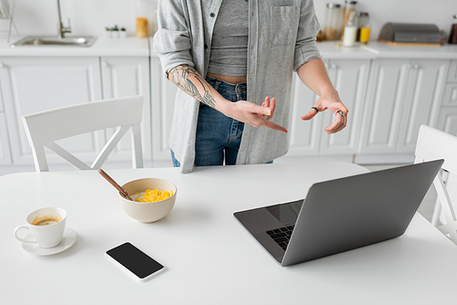 cropped view of young freelancer with tattoo on hand pointing at laptop near smartphone with blank screen, bowl with cornflakes, spoon and cup of coffee on white saucer on desk in modern kitchen