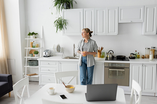 tattooed woman with bangs and eyeglasses holding glass of orange juice and standing near kitchen worktop next to desk with devices, bowl with cornflakes and cup of coffee on saucer at home, freelance