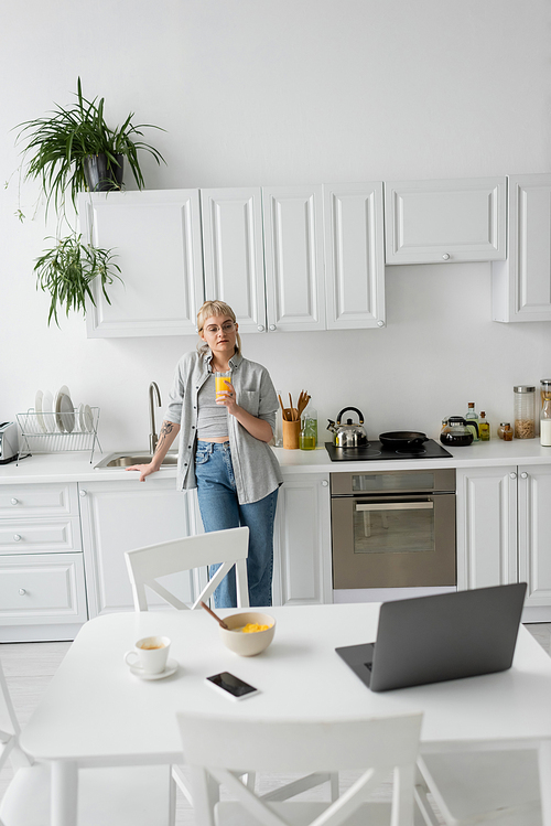tattooed woman in eyeglasses holding glass of orange juice and standing near desk with devices, bowl with cornflakes and cup of coffee with saucer on desk in modern kitchen, looking at laptop