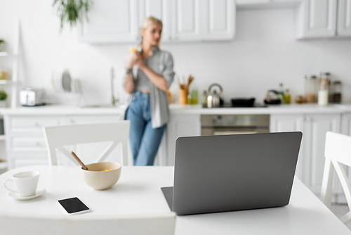 laptop, smartphone with blank screen, bowl with cornflakes and cup with saucer on white desk near blurred freelancer standing in modern kitchen, freelance lifestyle, digital nomad
