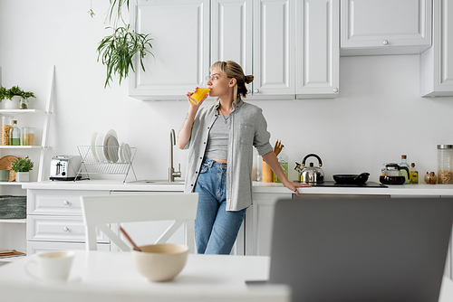 tattooed woman in eyeglasses drinking orange juice and standing near  kitchen worktop next to desk with devices, bowl with cornflakes and cup of coffee with saucer at home, freelance lifestyle