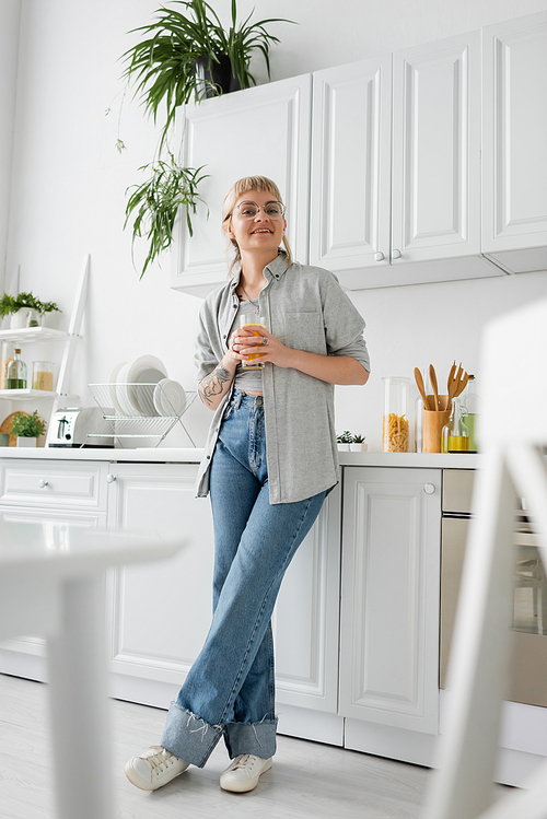 full length of tattooed and happy woman in eyeglasses holding glass of orange juice near kitchen worktop with clean dishes, toaster and rack with plants looking at camera in modern apartment