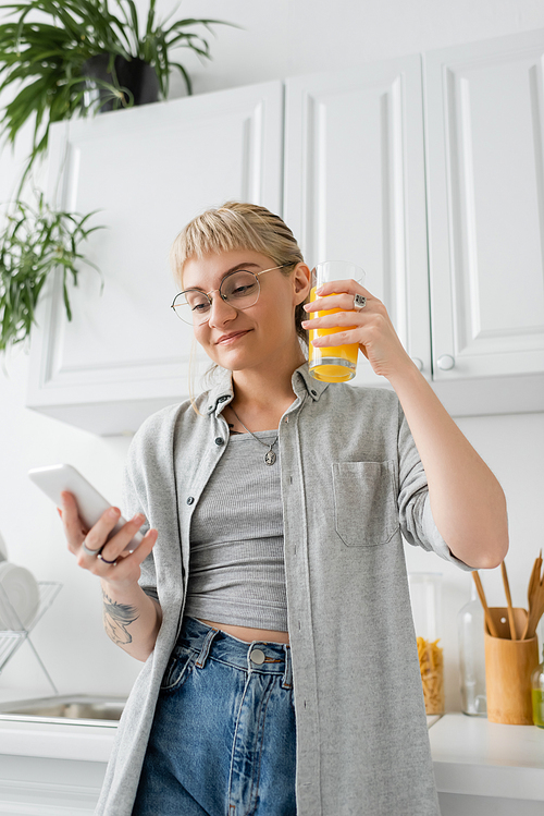 tattooed and happy woman with bangs and eyeglasses holding glass of orange juice and using smartphone while standing near clean dishes and blurred green plants in modern apartment