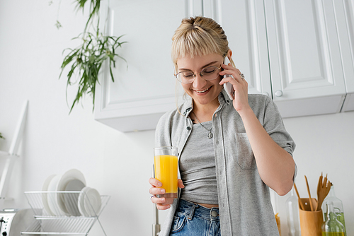 happy young woman with bangs and eyeglasses holding glass of orange juice and talking on smartphone standing with closed eyes in kitchen near blurred green plants in modern apartment