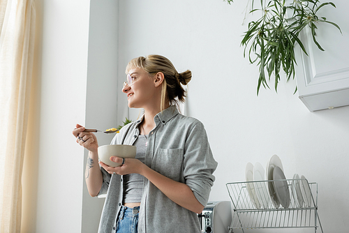 tattooed young woman with bangs and eyeglasses smiling while holding bowl with cornflakes and spoon while having breakfast and looking away near green plants and clean dishes in white kitchen