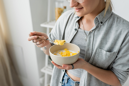 cropped view of tattooed young woman smiling while holding bowl with cornflakes and spoon while having breakfast next to blurred white rack in modern kitchen. copy space, apartment, morning energy