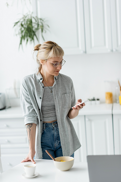 young woman with short hair, tattoo and bangs using smartphone while standing in eyeglasses near bowl with cornflakes, cup of coffee and laptop on table in modern apartment, freelancer
