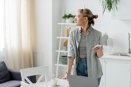 young woman with short hair, tattoo and bangs using smartphone while looking away and standing in eyeglasses near cup of coffee and blurred laptop on table in modern apartment, freelancer