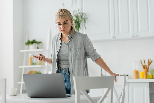 young woman with short hair, tattoo and bangs using smartphone while standing in eyeglasses near cup of coffee and looking at blurred laptop on table around chairs in modern apartment, freelancer