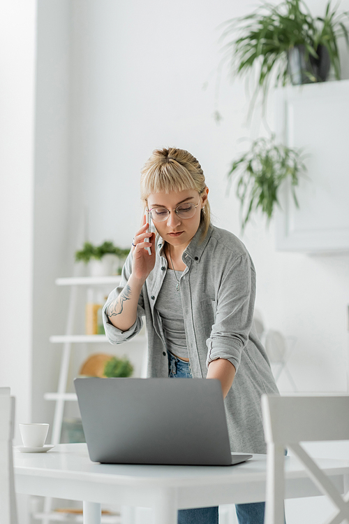 young woman with short hair, bangs and tattoo on hand talking on smartphone while standing in eyeglasses near cup of coffee and laptop on table in modern apartment, freelancer