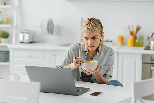 young woman with bangs and tattoo on hand eating cornflakes for breakfast while using laptop near smartphone with blank screen and cup of coffee on table in modern kitchen, freelancer