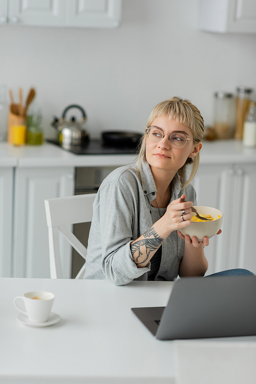 dreamy young woman with bangs, short hair and tattoo on hand eating cornflakes for breakfast while using laptop near cup of coffee with saucer on table in modern kitchen, freelancer, work from home