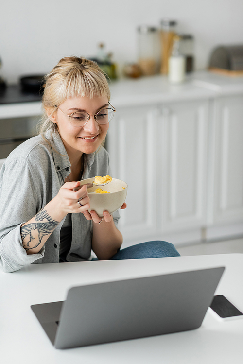 happy young woman with short hair, bangs and tattoo on hand eating cornflakes for breakfast while using laptop near smartphone with blank screen on table and smiling in modern kitchen, freelancer