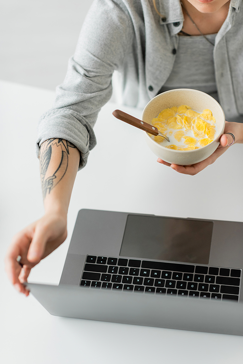 cropped view of young woman with tattoo on hand and grey shirt holding bowl with cornflakes with spoon while having breakfast and using laptop on white table in modern apartment, freelancer, top view