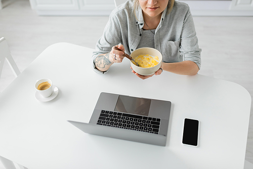 cropped view of young woman with tattoo on hand eating cornflakes for breakfast while using laptop near smartphone with blank screen and cup of coffee on table in modern kitchen, freelancer