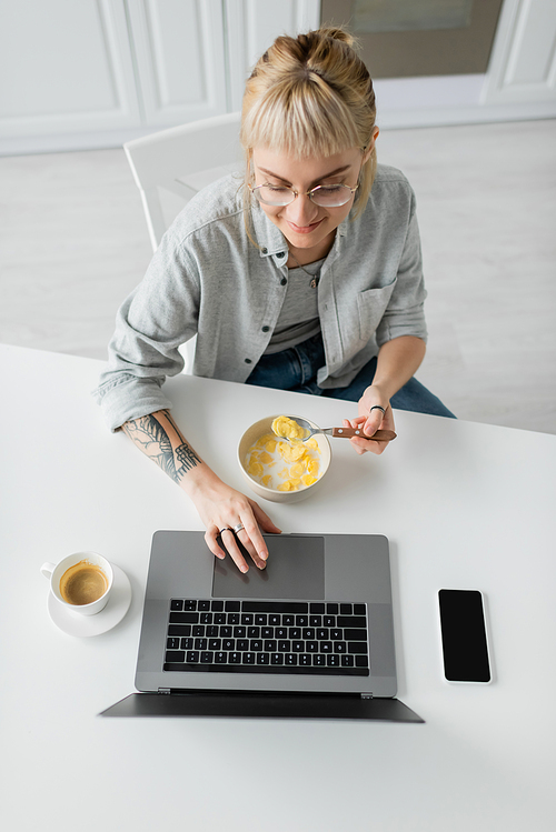 top view of happy young woman with tattoo on hand eating cornflakes for breakfast while using laptop near smartphone with blank screen and cup of coffee on table in modern kitchen, freelancer