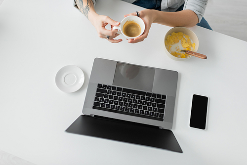 top view of woman with tattoo on hand holding cup of coffee near bowl with cornflakes during breakfast while using laptop near smartphone with blank screen in modern kitchen, freelancer, cropped