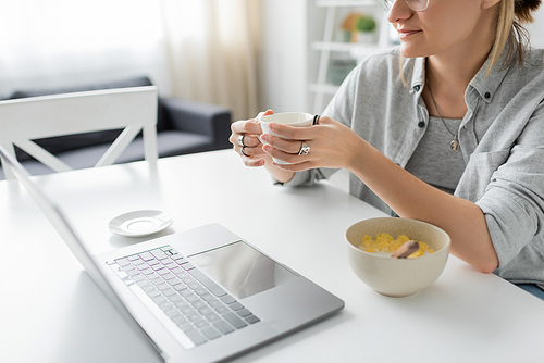 cropped shot of young woman holding cup of coffee near bowl with cornflakes during breakfast while using laptop near white saucer on table in modern kitchen, freelancer, work from home