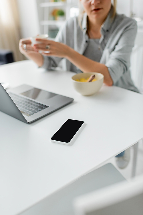 cropped shot of blurred woman holding cup of coffee near bowl with cornflakes during breakfast while using laptop near smartphone with blank screen in modern kitchen, freelancer