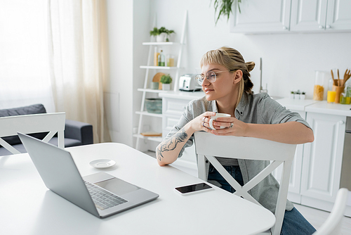 young woman with tattoo on hand and bangs holding cup of coffee and looking at laptop near smartphone and saucer on white table around chairs in modern kitchen, freelancer, remote lifestyle