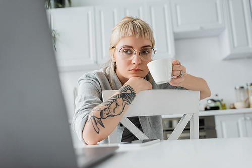 focused young woman with tattoo on hand and bangs holding cup of coffee and looking at blurred laptop near smartphone on white table in modern kitchen, freelancer, remote lifestyle