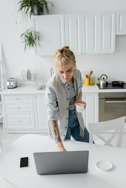 high anglve view of young woman with tattoo on hand and bangs holding cup of coffee and using laptop near smartphone and saucer on white table in modern kitchen, freelancer, remote lifestyle