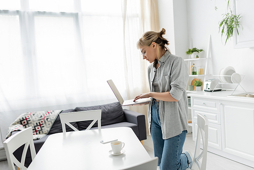 young woman in eyeglasses with short hair and bangs holding laptop near cup of coffee and smartphone on white table around chairs in white and modern kitchen next to sofa, freelancer, remote lifestyle