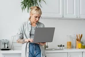 tattooed young woman in eyeglasses, short hair and bangs holding and using laptop while working from home in white and modern kitchen, blurred background, remote lifestyle, freelancer