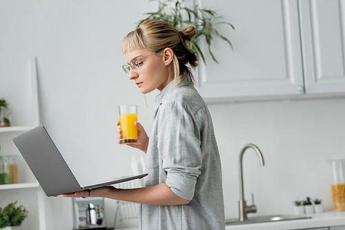young woman in eyeglasses, short hair and bangs holding glass of fresh orange juice and using laptop while working from home in white and modern kitchen, blurred background, freelancer