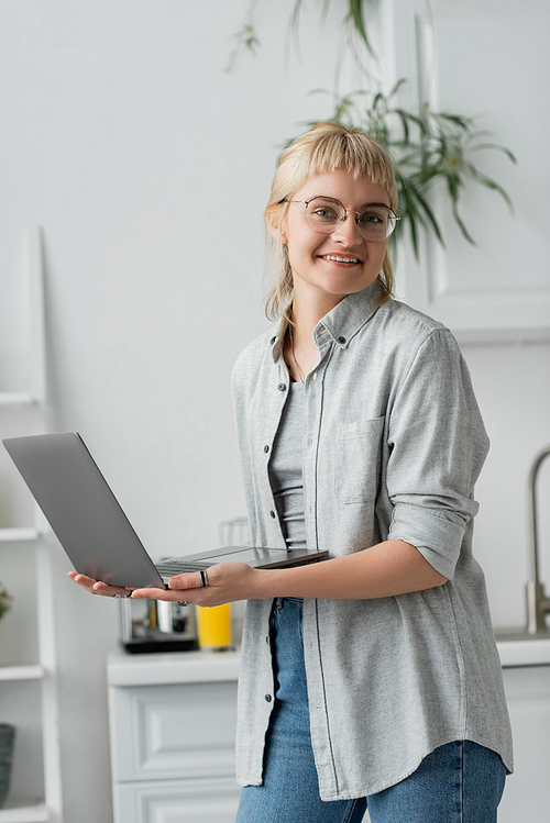 happy young woman in eyeglasses, short hair and bangs holding and using laptop while looking at camera and working from home in modern kitchen, blurred background, remote lifestyle, freelancer