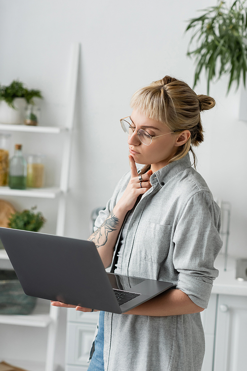 pensive young woman in eyeglasses, with tattoo and bangs using laptop while working from home in white and modern kitchen, blurred background, remote lifestyle, freelancer, hand near chin