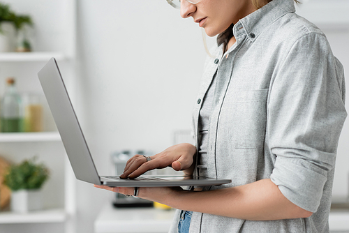 cropped view of focused young woman in grey shirt holding and using laptop in white and modern kitchen, blurred background, remote lifestyle, freelancer, work from home, self-employed