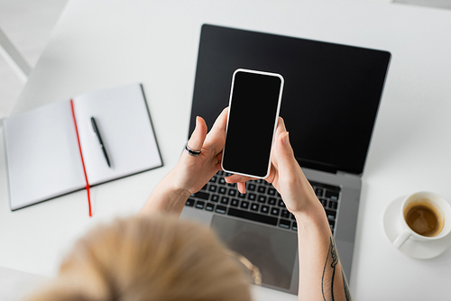 top view of tattooed woman holding smartphone with blank screen near laptop, notebook with pen, and cup of coffee with saucer on white table while working from home, freelancer, modern workspace