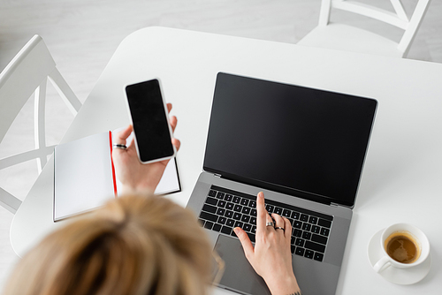 top view of blurred woman holding smartphone with blank screen near laptop, notebook with pen, and cup of coffee with saucer on white table while working from home, freelancer, modern workspace