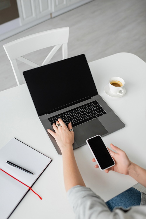 top view of woman holding smartphone with blank screen and using laptop near notebook with pen, and cup of coffee with saucer on white table while working from home, freelancer, modern workspace