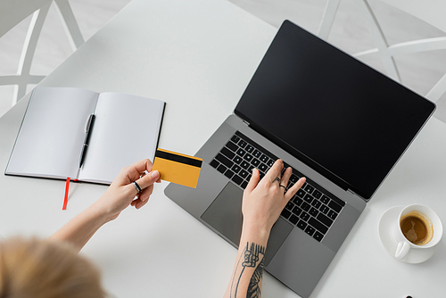 top view of tattooed young woman holding credit card and using laptop with blank screen near notebook with pen, and cup of coffee with saucer on white table, modern workspace, work from home